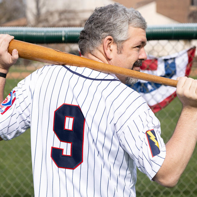 A man with short gray hair and a beard is seen from behind, wearing a white pinstriped baseball jersey with the number "9" on the back. He holds a wooden baseball bat over his shoulders with both hands. The jersey features a patch on the sleeve with a lightning bolt emblem. He is standing near a chain-link fence, with a red, white, and blue bunting decoration hanging in the background. The setting is an outdoor baseball field with grass and buildings visible beyond the fence.