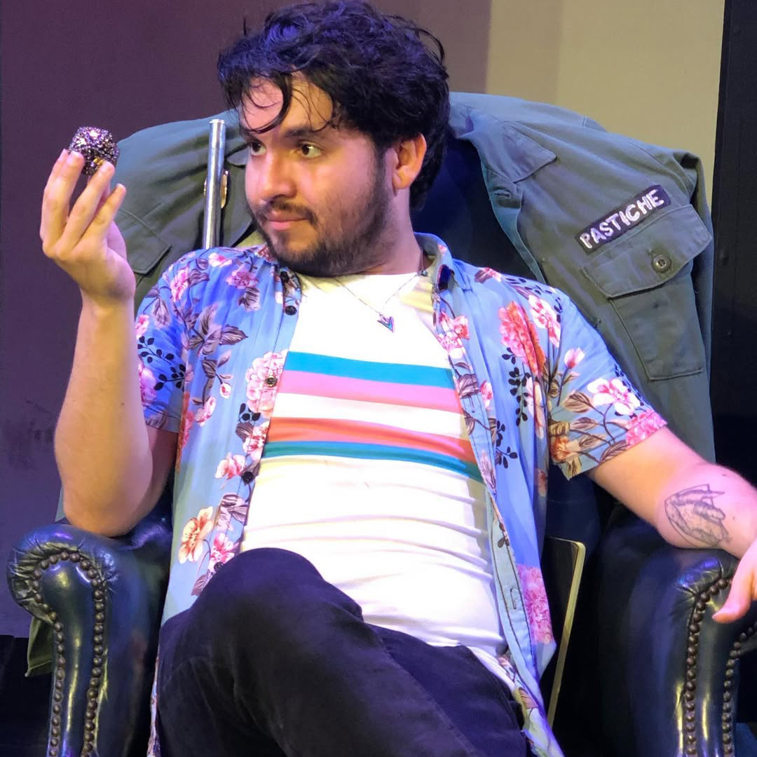 A young person with dark, wavy hair and a beard sits in a green leather armchair, holding a small, glittery object in their right hand while gazing at it with a thoughtful expression. They wear a floral Hawaiian-style shirt over a striped transexual pride t-shirt, and a green jacket with a patch reading "PASTICHE" is draped over the back of the chair. Their left arm rests on the armrest, revealing a tattoo on their forearm. The background is dimly lit, with a stage-like setting.