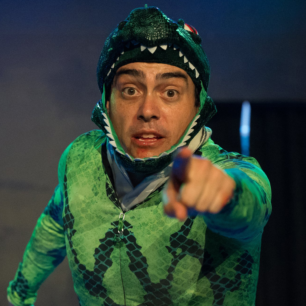 A man wearing a green, scaly-patterned lizard costume with a hood resembling a lizard head is pointing directly at the camera. His facial expression is intense, with wide eyes and an open mouth. The background is dimly lit with blue lighting.